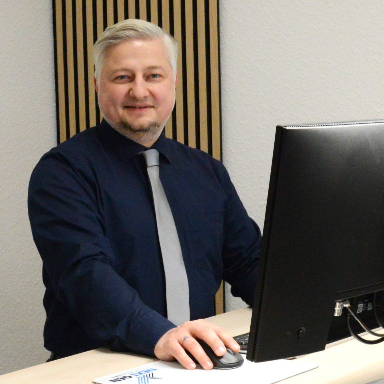 Mann in einem dunkelblauen Hemd und mit einer Krawatte steht vor einem Computer.