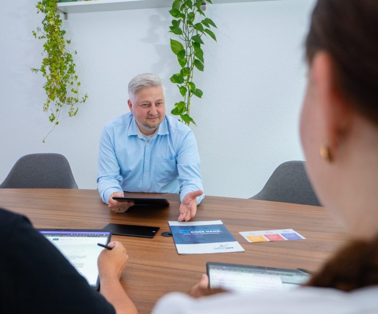 Mann im blauen Hemd sitzt am Tisch und spricht mit zwei Personen. Pflanzen im Hintergrund.