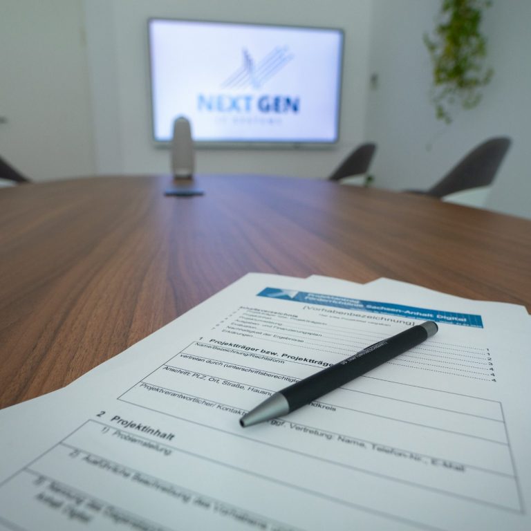 Ein Tisch mit Dokumenten und einem Stift, im Hintergrund ein Bildschirm mit NextGen.