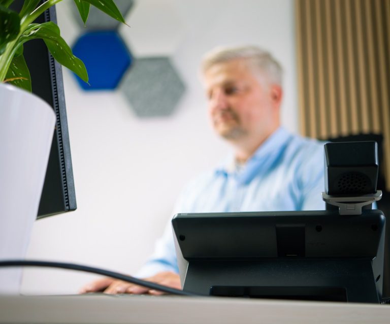 Mann im blauen Hemd sitzt am Tisch und spricht mit zwei Personen. Pflanzen im Hintergrund.
