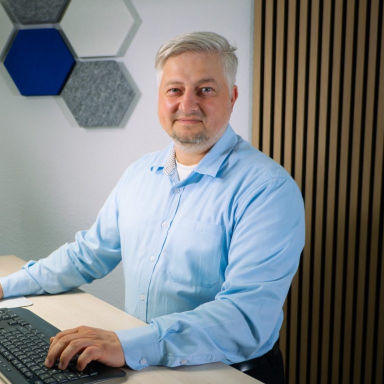 Mann mit blauer Hemd steht an einem Schreibtisch mit Computer und lächelt.